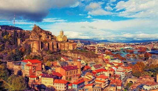 Tbilisi skyline with historic Narikala Fortress on hilltop overlooking colorful old town buildings and distant mountains unde
